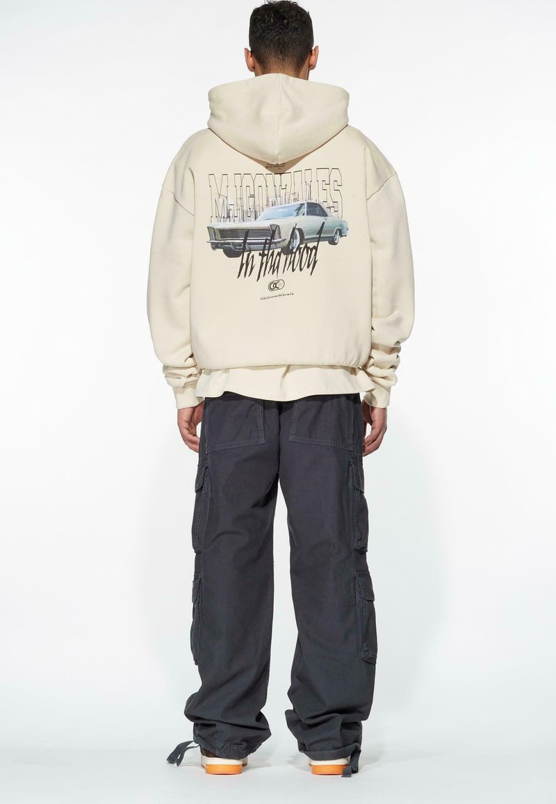 Beige Kapuzenpullover mit Rückendruck eines klassischen Autos, kombiniert mit dunklen Cargo-Hosen mit mehreren Taschen und elastischen Bündchen, getragen mit orange-soled Schuhen.
