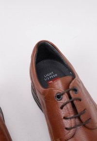 Brown leather shoe featuring a smooth texture, rounded toe, subtle stitching details, and black laces. Interior has a gray lining and label, "LIGHT 1/2 PESO."