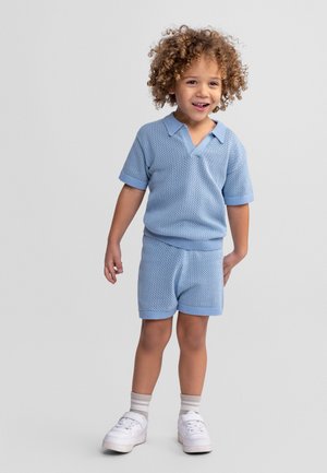 Enfant souriant aux cheveux bouclés portant un haut à manches courtes tricoté bleu clair et un short assorti, des chaussettes blanches et des baskets blanches.