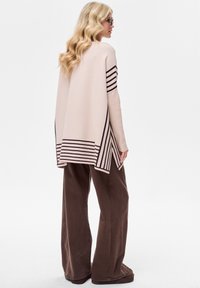 Lichtroze gebreide trui met diagonale bruine strepen, gecombineerd met bruine broek met wijde pijpen. De trui heeft een slappere pasvorm en een langere achterkant.