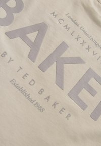 Béžová tkanina s veľkými textúrovanými šedými písmenami. "BAKER" je výrazne zobrazené, s menším textom uvádzajúcim značku a rok založenia.