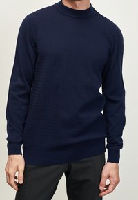 Man in een donkerblauwe gebreide trui met lange mouwen en zwarte broek tegen een effen achtergrond.