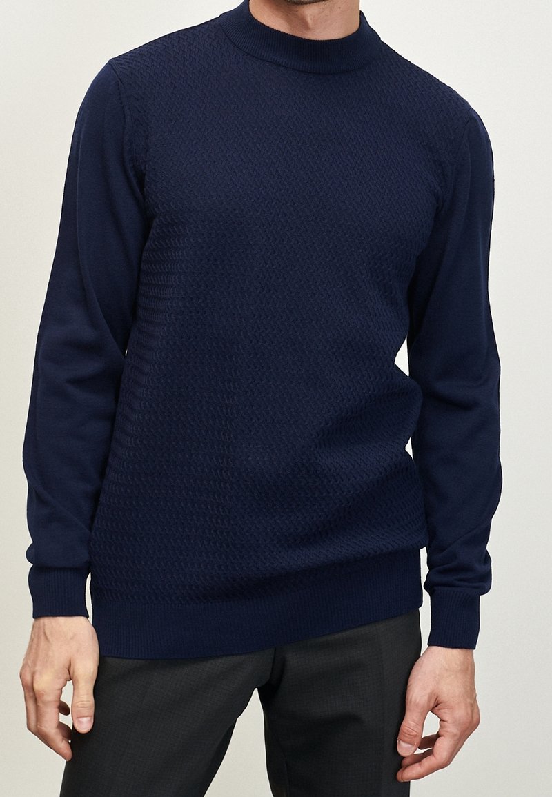 Man in een donkerblauwe gebreide trui met lange mouwen en zwarte broek tegen een effen achtergrond.
