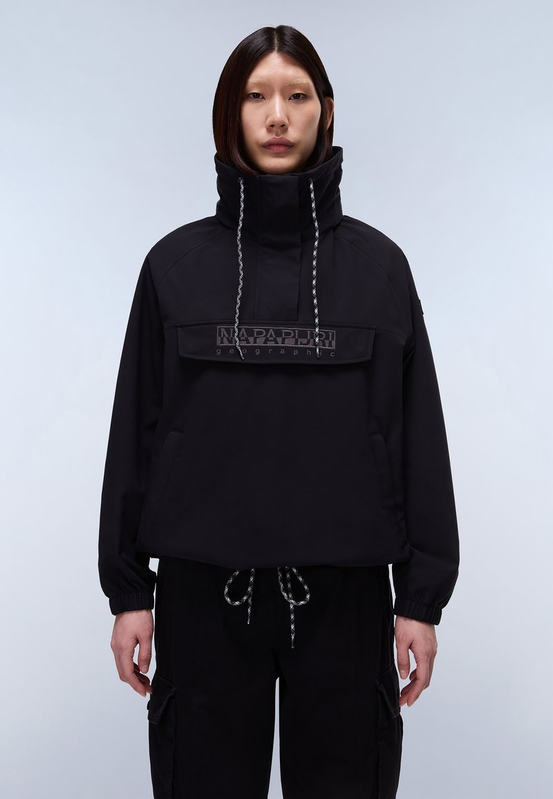 Άτομο με ίσια σκούρα μαλλιά φοράει μαύρο πουλόβερ με ψηλό γιακά, τσέπη με λογότυπο μπροστά και κορδόνια, μαύρο παντελόνι με πλαϊνές τσέπες τύπου cargo.