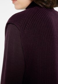 Gestickte Weste in Dunkellila, mit einem strukturierten Muster auf den Schultern, kombiniert mit einem glatten weinroten Langarmshirt darunter.