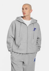 Szara bluza z kapturem i suwakem, z niebieskim logo na piersi. Materiał wydaje się miękki, a krój jest luźny.