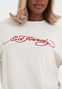 Kremfarvet sweatshirt med rødt broderet "Ed Hardy" logo. Blød tekstur med en afslappet pasform og rund halsudskæring.