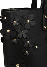 Černá kožená taška s texturovanými černými květinovými aplikacemi a doplňky ve zlaté barvě. Obsahuje dvě rukojeti pro nošení.