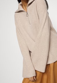 Beiger, gerippter Strickpullover mit hohem Kragen und halbem Reißverschluss. Verfügt über breite Ärmel und eine weiche Textur, getragen über einem beigen Rock.