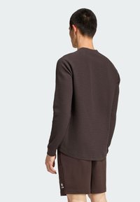 Tmavo hnedá košeľa s dlhým rukávom z textúrovanej látky, s rebrovanými manžetami a zaobleným lemom, v kombinácii s rovnako farbenými šortkami s bočnými vreckami.