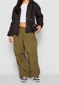 Gesteppte schwarze Jacke mit Kapuze, weißes Croptop, olivgrüne, weit geschnittene Hosen und schwarze Umhängetasche. Sneakers sind schwarz-weiß.