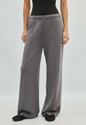 Personne portant un pantalon de jogging gris ample avec une taille à cordon et des chaussures noires à enfiler, debout devant un fond clair uni.