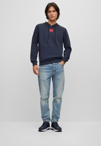 Marineblå hettegenser med frontlomme og rød logo, kombinert med lyseblå slitte jeans og marineblå joggesko. Enkelt, uformelt design.