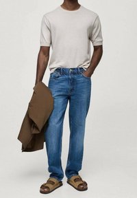 Light grey short-sleeve t-shirt, blue straight-leg jeans, brown jacket draped over arm, and tan sandals with straps.