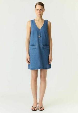 Kvinde står med fronten frem iført en ærmeløs blå denimkjole med påsyede lommer, en lang halskæde og sorte sandaler mod en ensfarvet baggrund.