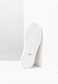 Zapatilla de ante beige con suela de goma blanca texturizada, que presenta un patrón ondulado elevado y detalles de marca en un inserto rectangular. Talla 37.