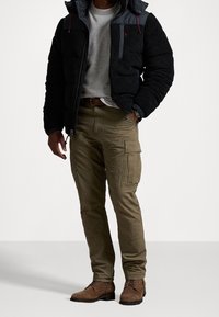 Man i svart fleecejacka, grå sweatshirt, olivgrön cargo-byxor, brun bälte, armbandsur och bruna mockastövlar står med en hand i fickan.