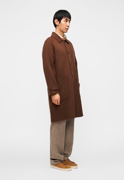 Καφέ παλτό από μάλλινο ύφασμα με κουμπιά μπροστά, μυτερό γιακά και δύο μπροστινές τσέπες, συνδυασμένο με καφέ υφασμάτινο παντελόνι με υφή και ταμπά αθλητικά παπούτσια.