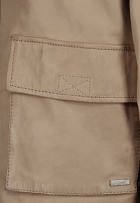 Beige läderjacka med en fackficka, synliga sömmar och en silverfärgad logotypetikett, som framhäver den släta texturen.