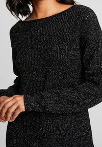 Nainen, joka pukeutunut mustaan teksturoituun neulepaitaan, jossa on pitkät hihat ja hopealankadetaljit, kädet kevyesti yhteen kiedottuina edessä.