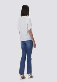 Blusa bianca con pizzo sulle maniche corte, abbinata a jeans blu aderenti e scarpe con tacco argento, vista da dietro.