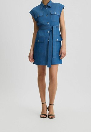 Kobieta w niebieskiej, bezrękawnikowej sukience dżinsowej z paskiem, guzikami i przednimi kieszeniami, w czarnych szpilkach z paskiem wokół kostki, stojąca na szarej podłodze.