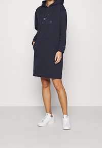 Vestito felpa con cappuccio blu navy con logo glitterato, tasca a marsupio, polsini a costine e vestibilità comoda, abbinato a sneakers bianche.