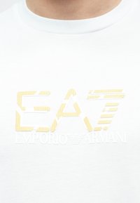 Biele tričko s okrúhlym výstrihom s béžovými textúrovanými písmenami "EA7" a menším bielym nápisom "Emporio Armani" pod nimi na hrudi.