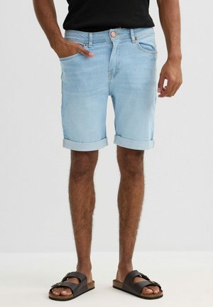 Hombre con pantalones cortos de mezclilla azul claro remangados, camiseta negra y sandalias marrón oscuro con hebillas, de pie con una mano en el bolsillo.