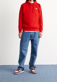 Röd Nike-hoodie med framlomma, blå denimbyxor och maroon- och vita sneakers med svarta detaljer. Slät tyg och avslappnad passform.