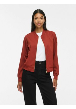 Femme portant une veste bomber zippée couleur rouille sur une chemise blanche avec un pantalon noir taille haute, sur un fond blanc uni.