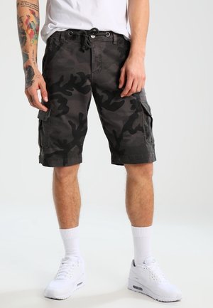 Ein Mann trägt dunkelgraue und schwarze Tarn-Cargoshorts, weiße Socken bis zur Wade und weiße Sneakers. Er steht vor einem schlichten Hintergrund.