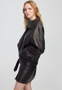 Giacca di pelle nera con vita a cintura, vestibilità oversize e colletto alto, con una texture liscia e un design minimalista.