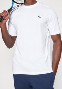 Vit t-shirt med kort ärm i bomull, rund halsringning och en grön Lacoste-logotyp på bröstet, hålls av en person med en tennisracket.