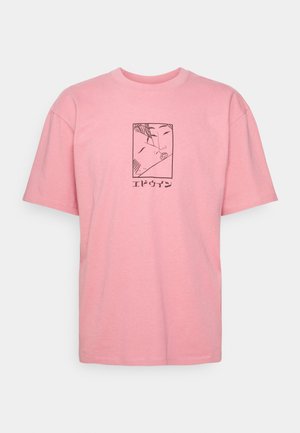 Mies, jolla on päällään vaaleanpunainen kuvioitu t-paita, mustat shortsit, mustat sukat, sinapinkeltaiset lenkkarit ja joka pitää yhdessä kädessä mustaa baseball-lippistä.