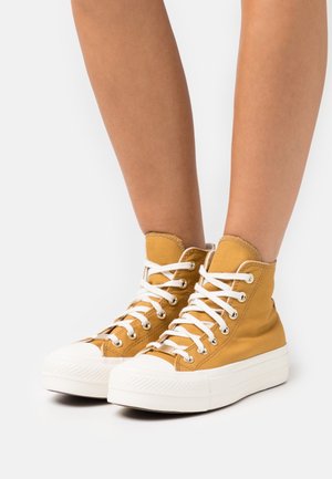 Benedenbenen met mosterdgele hoge canvas sneakers met witte veters en dikke witte rubberen zolen op een witte achtergrond.