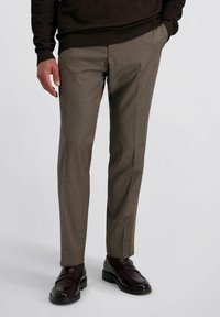 Man in slim-fit taupe pantalon met een hand in zijn zak en donkerbruine leren loafers op een witte achtergrond.