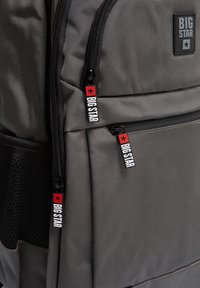 Sivi ruksak s teksturiranim materijalom, crnim patentnim zatvaračima s logotipom na povlačenju i mrežastom bočnom džepu. Paket s logotipom na prednjoj strani.