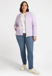 Giacca imbottita viola chiaro con texture in tessuto trapuntato, chiusura con zip e due tasche frontali. Abbinata a un top bianco, jeans blu e scarpe chiare.