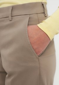 Μπεζ παντελόνι με ομαλή υφή διαθέτει πλαϊνή τσέπη και ραμμένες ραφές. Ένα κίτρινο πλεκτό πουκάμισο με μακριά μανίκια είναι μερικώς ορατό.