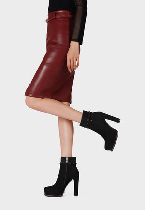 Donna che indossa una gonna in pelle bordeaux, un top nero trasparente a maniche lunghe e stivaletti neri con tacco alto e dettagli con bottoni.