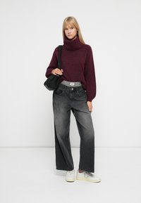 Maglione a collo alto bordeaux, jeans grigi a gamba larga e sneaker bianche. Una borsa a spalla nera e una cintura a fantasia completano l'outfit.