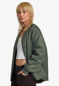 Prešívaná bomber bunda v olivovo zelenej farbe, s okrúhlym krkom, gombíkovým zapínaním a bočnými vreckami. Nosená cez biele cropped tričko.