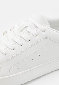 Sneaker in pelle bianca con punta tonda, suola piatta in gomma e dettagli perforati sui lati; lacci infilati attraverso gli occhielli.