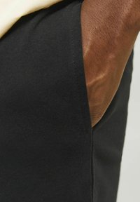 Tessuto nero con texture a coste, presentato con una tasca visibile e una manica parziale. Il materiale appare morbido e aderente intorno alla silhouette.
