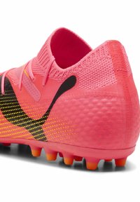 Bota de fútbol de un brillante color rosa con parte superior de punto, diseño de acentos en negro y patrón de puntos amarillos; presenta tacos en naranja y negro en la suela.