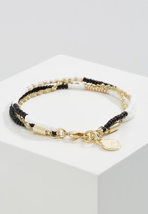 Bracciale multi-filo con perline nere, bianche e dorate, maglie di catena dorate, chiusura a moschettone e un piccolo ciondolo rotondo dorato su una superficie bianca.