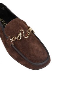 Brązowe zamszowe mokasyny z złotym łańcuchem jako akcentem i zaokrąglonym noskiem. Smukły design z kontrastującym ciemnym wykończeniem wzdłuż krawędzi buta.