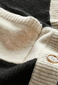 Knitted cardigan with segments of cream, beige, and black fabric. Features a ribbed texture and a large round button.