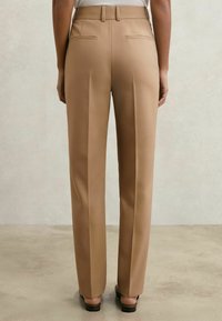 Vedere din spate a unei persoane care poartă pantaloni bej mulați, cu croială dreaptă și pantofi negri cu toc, stând în picioare pe un fundal simplu și neutru.